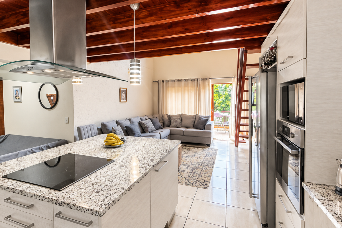 Spacious open-plan living area featuring a modern kitchen with granite countertops, built-in appliances, and striking exposed timber ceiling beams leading to a balcony.