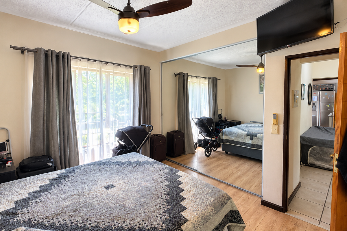 A bedroom featuring laminate flooring, a ceiling fan, and large mirrored sliding wardrobe doors, with an adjacent view into a tiled hallway.