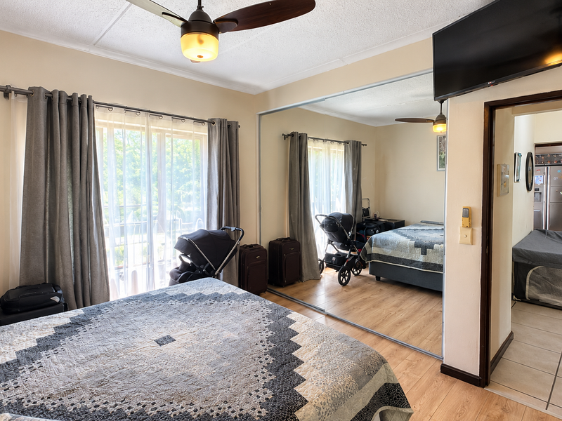 A bedroom featuring laminate flooring, a ceiling fan, and large mirrored sliding wardrobe doors, with an adjacent view into a tiled hallway.
