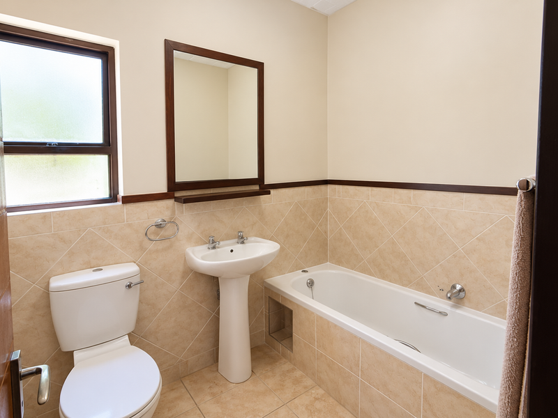 A clean and functional bathroom featuring a pedestal basin, bathtub, toilet, and neutral wall tiling with a wooden-framed mirror.
