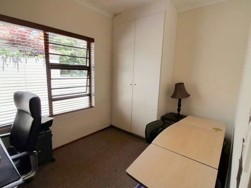 A functional home office featuring neutral walls, carpet flooring, a built-in cupboard for storage, and a window with horizontal blinds.