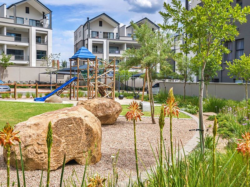 A well-maintained communal garden area featuring landscaped beds, large decorative boulders, and a children's playground set within an apartment complex.
