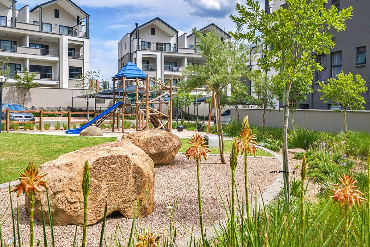A well-maintained communal garden area featuring landscaped beds, large decorative boulders, and a children's playground set within an apartment complex.