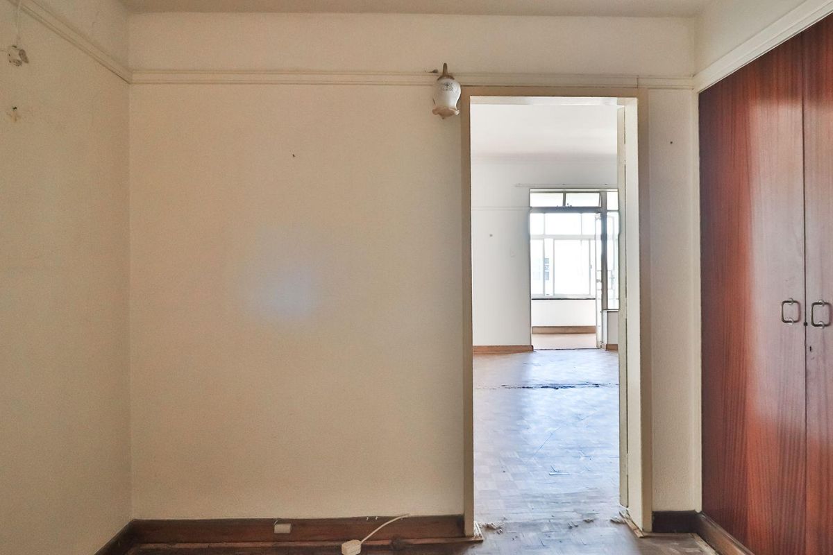 An interior view of a bedroom featuring built-in wooden cupboards and parquet flooring, leading through a doorway into an adjacent living area.
