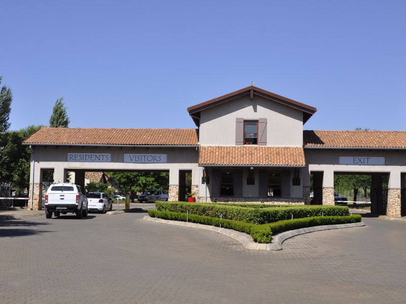 A well-maintained residential estate entrance featuring a security gatehouse with dedicated resident and visitor lanes, paved driveway, and manicured landscaping.