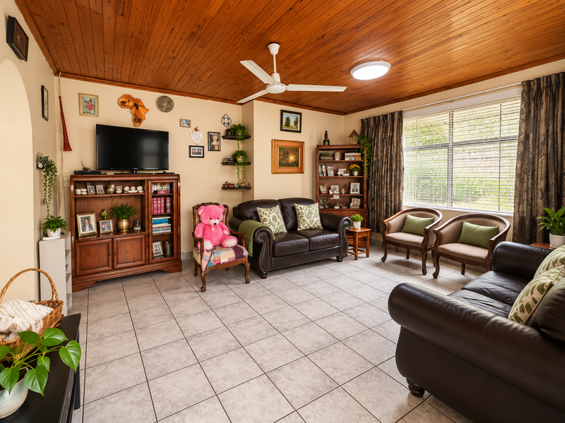 A comfortable living room featuring a warm timber tongue and groove ceiling, ceiling fan, and neutral tiled flooring with large window for natural light.