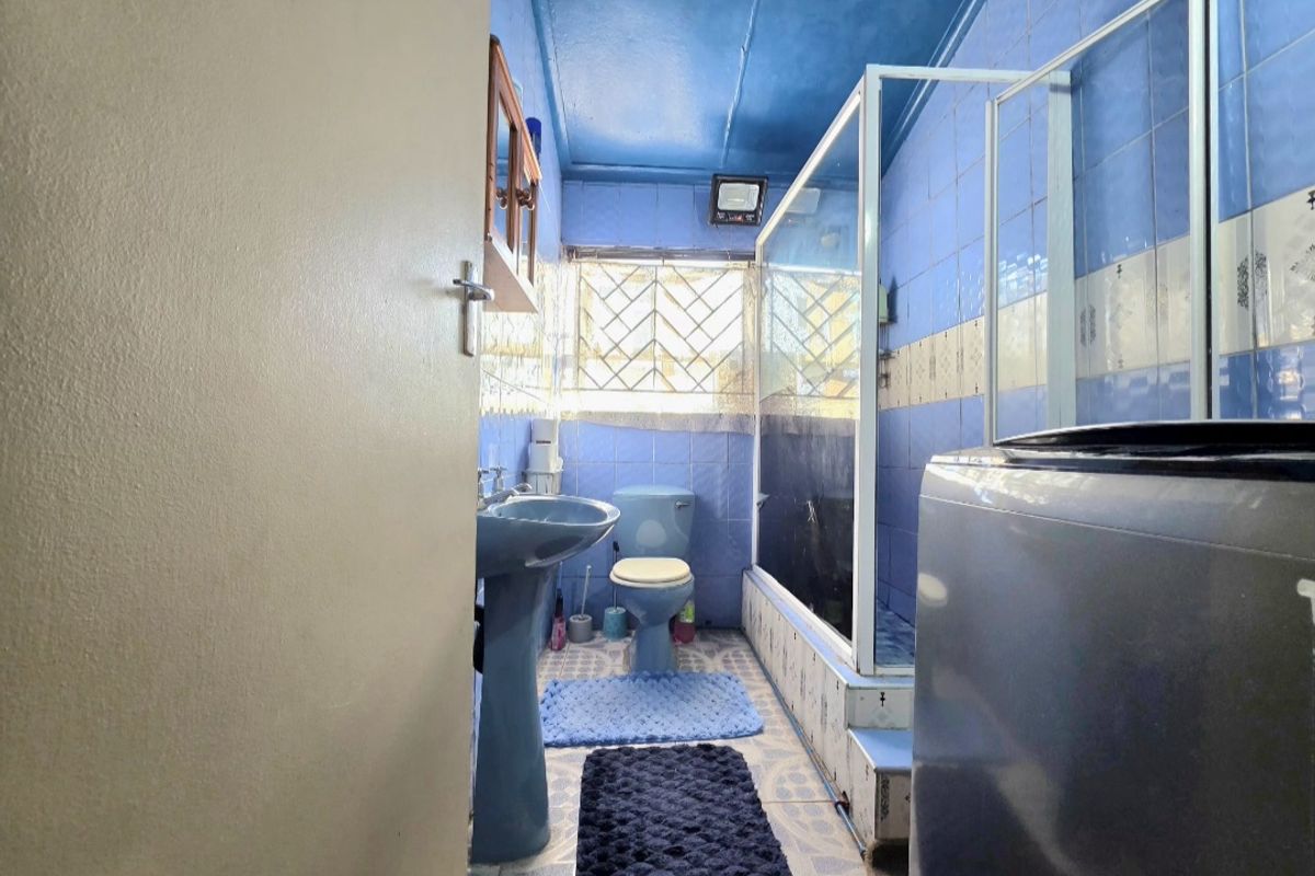 A compact bathroom featuring blue wall tiles, a pedestal basin, toilet, and a shower enclosure with security bars on the window.
