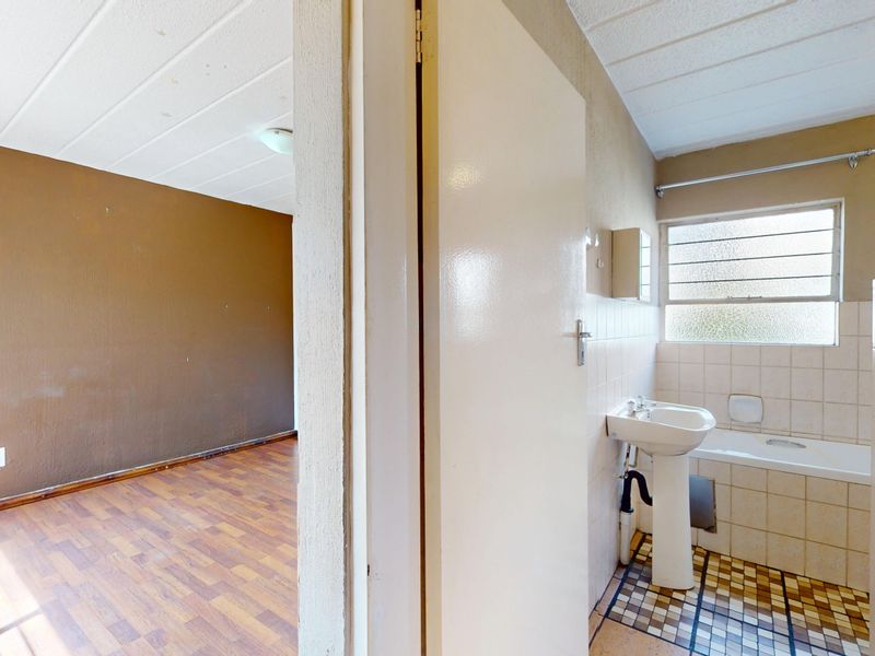 A dated bathroom featuring a pedestal basin and bathtub, adjacent to an empty room with wood-look flooring.