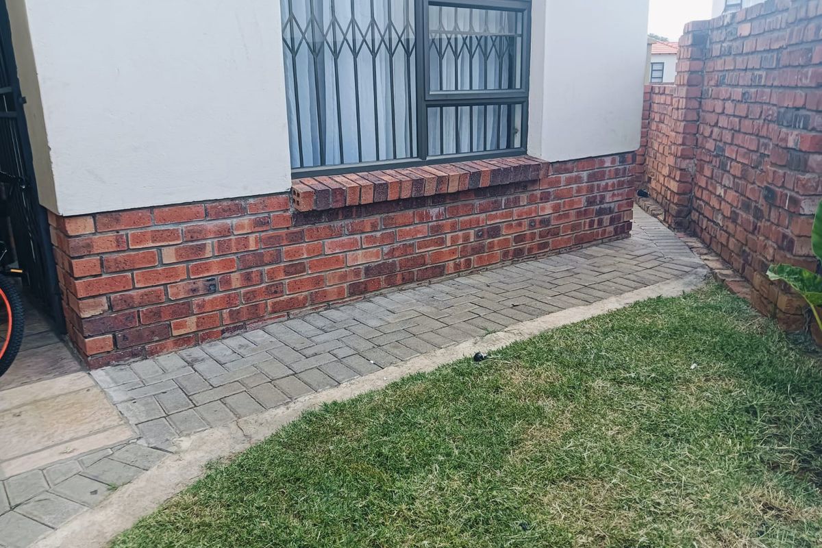 Rear exterior view of the townhouse featuring a paved walkway, brick boundary wall, and security bars on the window.