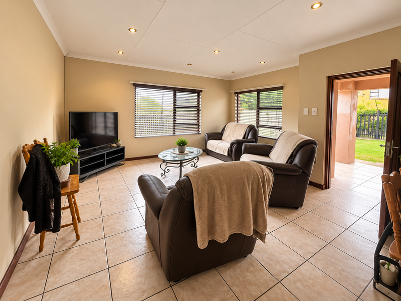 A well-lit living room featuring neutral tiled flooring, recessed ceiling lighting, and large windows with venetian blinds, providing direct access to an outdoor patio area.
