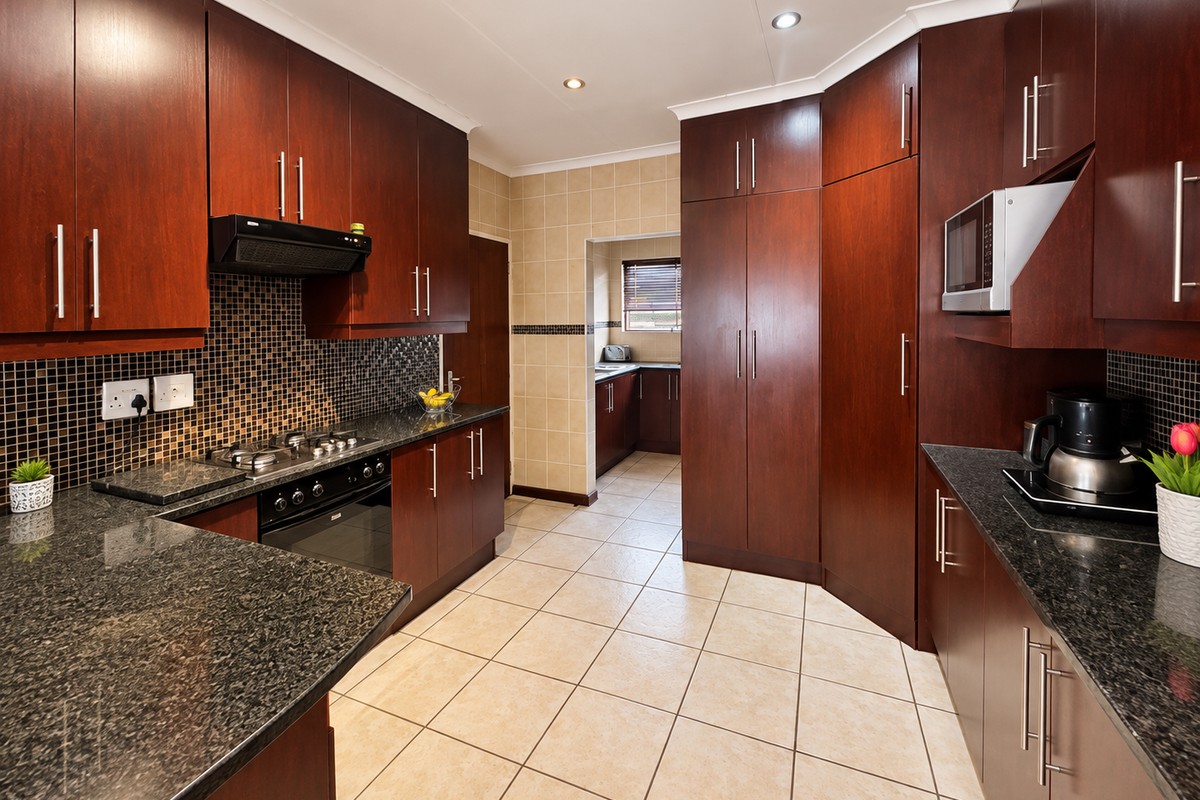 A well-maintained kitchen featuring dark wood cabinetry, granite countertops, a gas hob, built-in oven, and a tiled backsplash, with an adjoining scullery area.