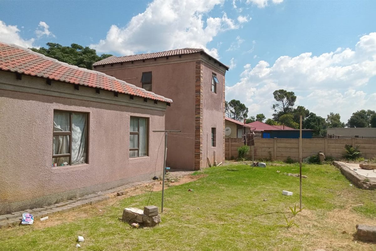 Rear view of the property featuring a multi-level structure with tiled roofing, plastered walls, and a spacious backyard with boundary walling.