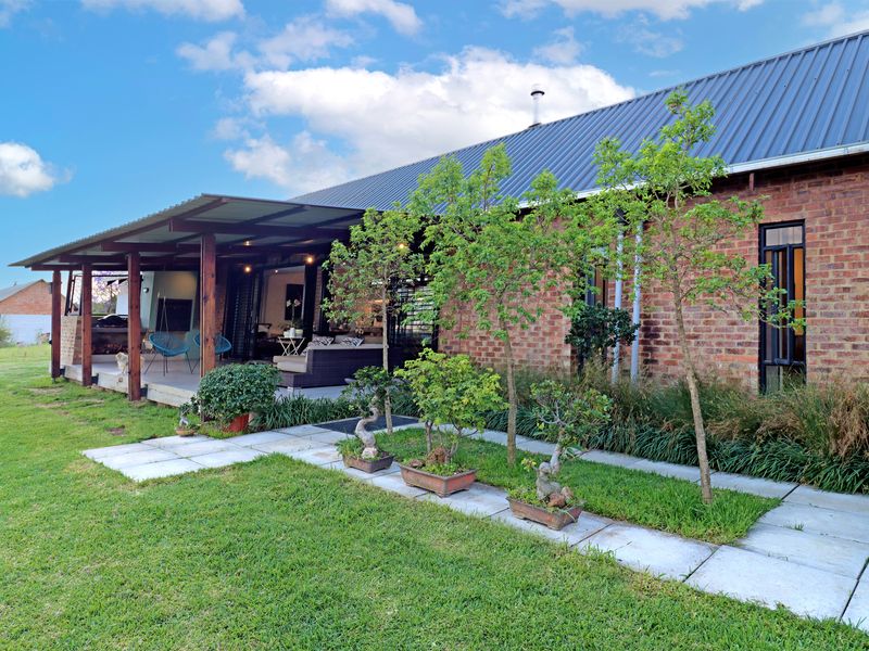Rear view of a brick house featuring a covered patio with timber beams, a built-in braai, and a well-maintained garden with paved pathways.