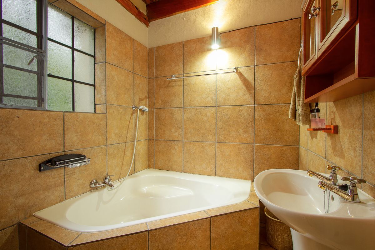 A compact bathroom featuring a corner bathtub, pedestal basin, and full-height wall tiling with exposed timber ceiling beams.