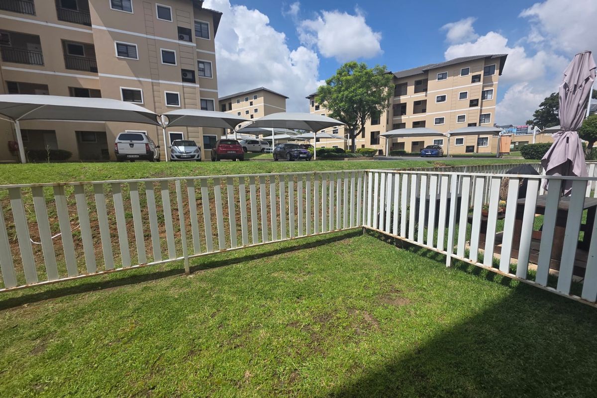A private rear garden area featuring a lawn and metal perimeter fencing, overlooking the apartment complex grounds and communal carport structures.