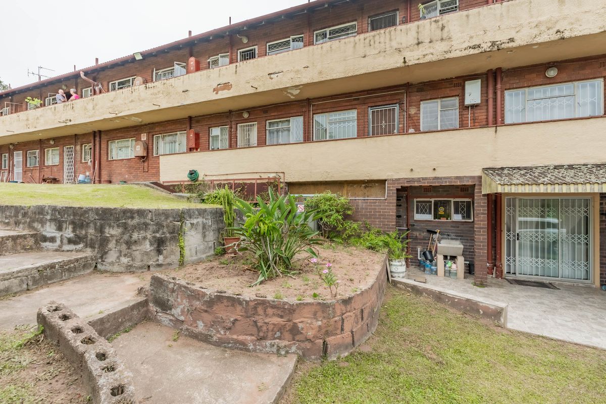 Rear view of a multi-story apartment building featuring brick construction, concrete balconies with visible wear, and a small garden area with a retaining wall.