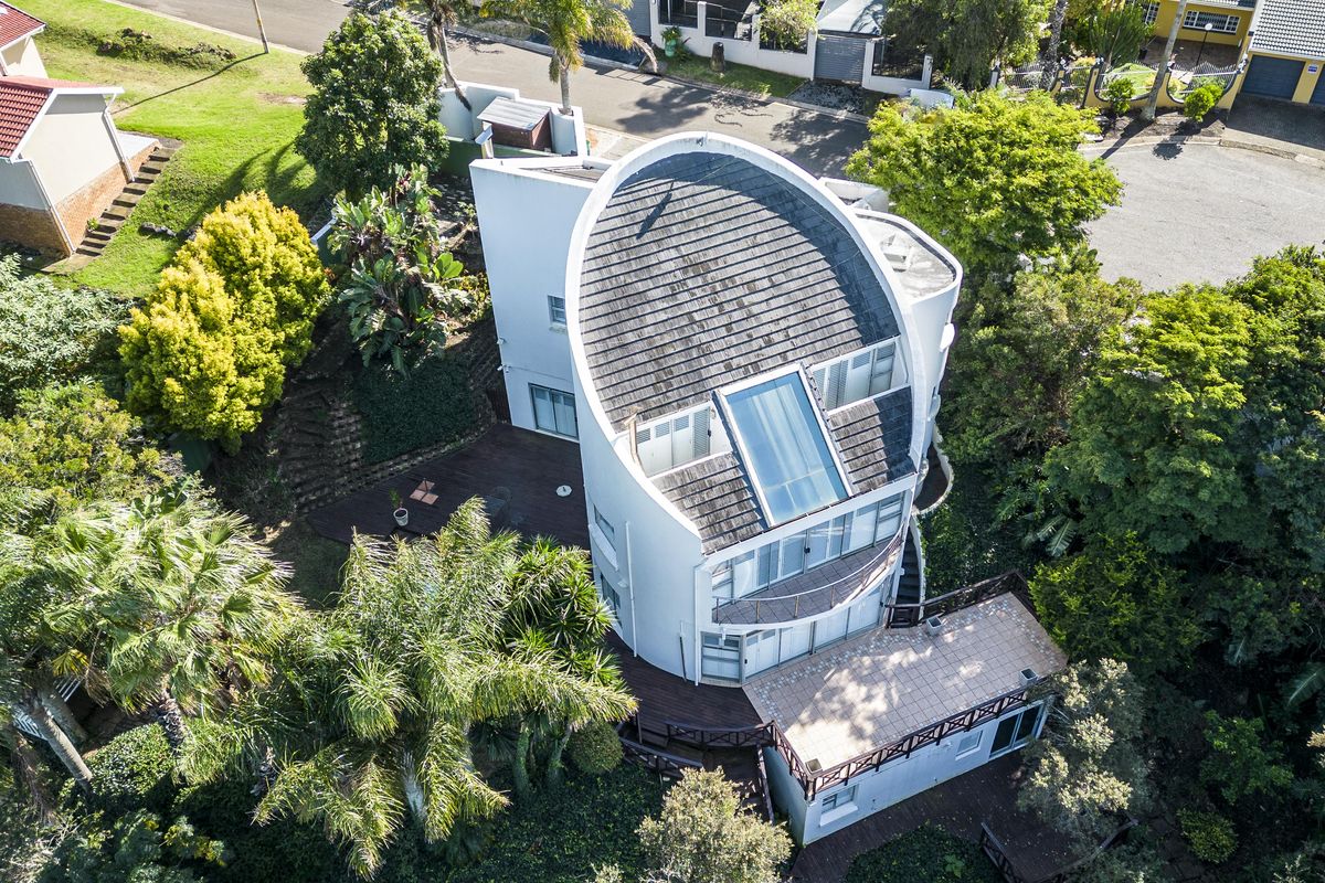 Aerial view of a unique architectural home featuring a distinctive curved roof, integrated skylight, expansive timber decking, and established surrounding gardens.