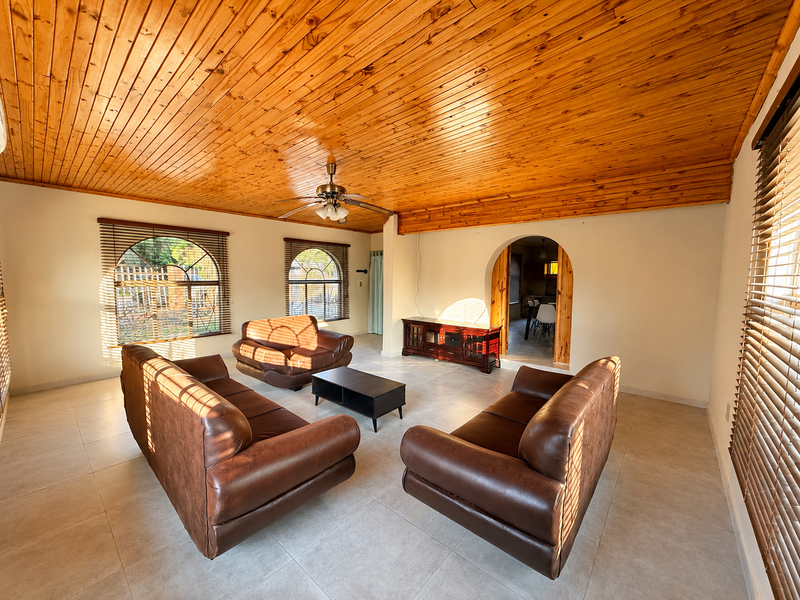 Spacious living room featuring a warm timber tongue and groove ceiling, tiled flooring, and arched windows with venetian blinds, leading to an adjacent room.