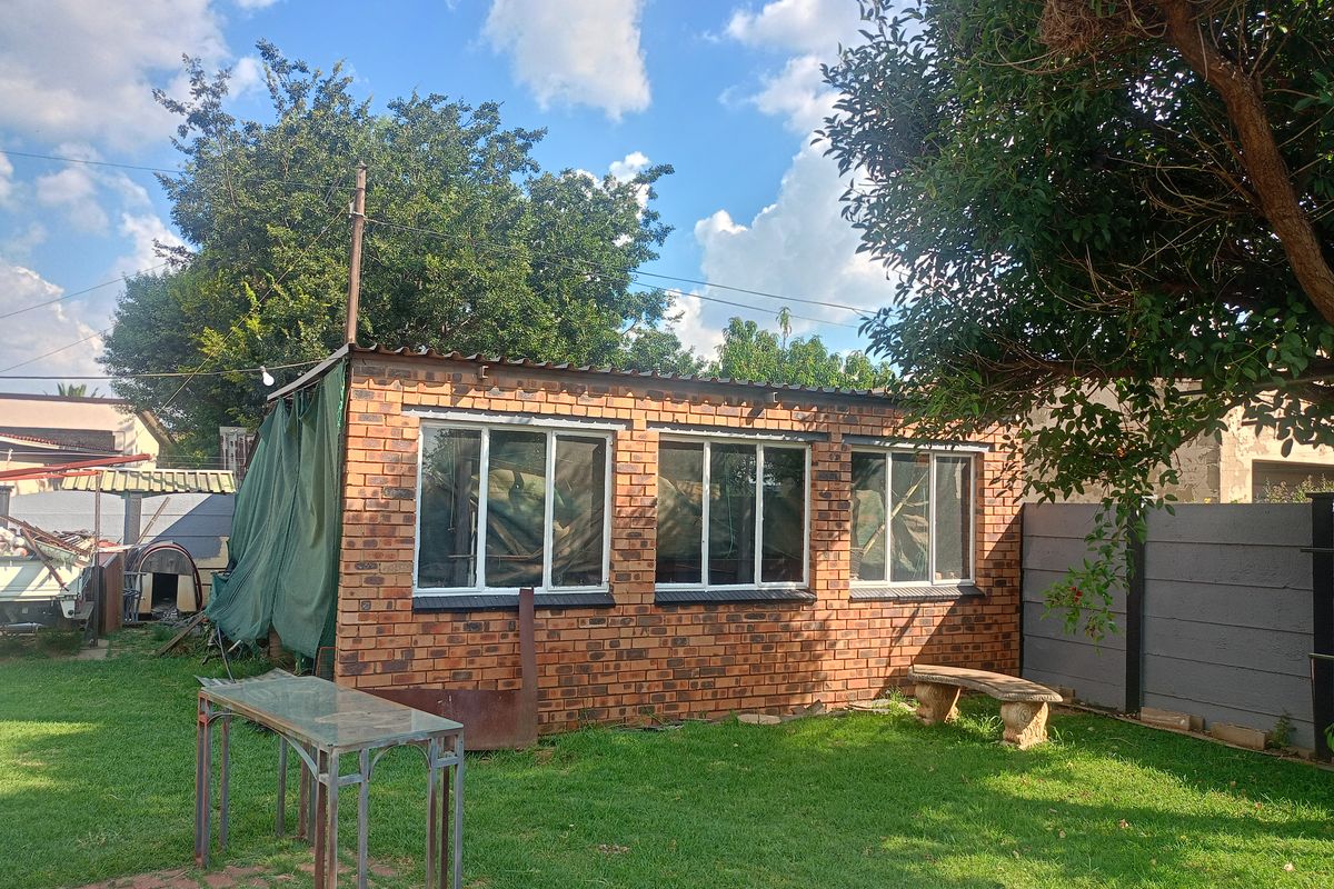 A detached brick outbuilding situated in a garden setting, featuring multiple windows and a corrugated roof structure.