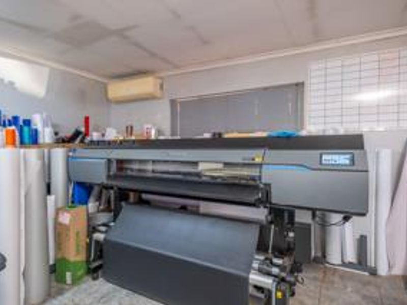 A utility or workshop space featuring a wall-mounted air conditioning unit and tiled wall sections, currently utilized for commercial printing equipment.