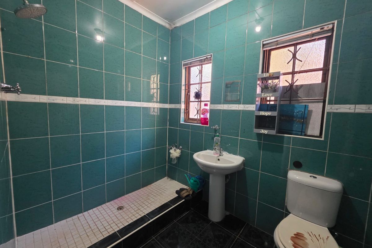 A functional bathroom featuring a pedestal sink, toilet, and a tiled shower area with dated green wall tiles and security bars on the windows.