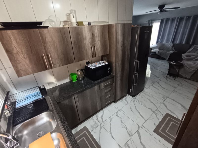 Open-plan kitchen featuring modern wood-look cabinetry, granite countertops, and a double basin sink, flowing seamlessly into the adjacent living area with tiled floors.