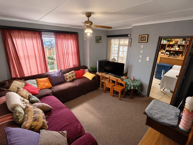 A comfortable living room featuring a ceiling fan, coved cornicing, and window security bars, with an adjacent view into a secondary utility or storage area.