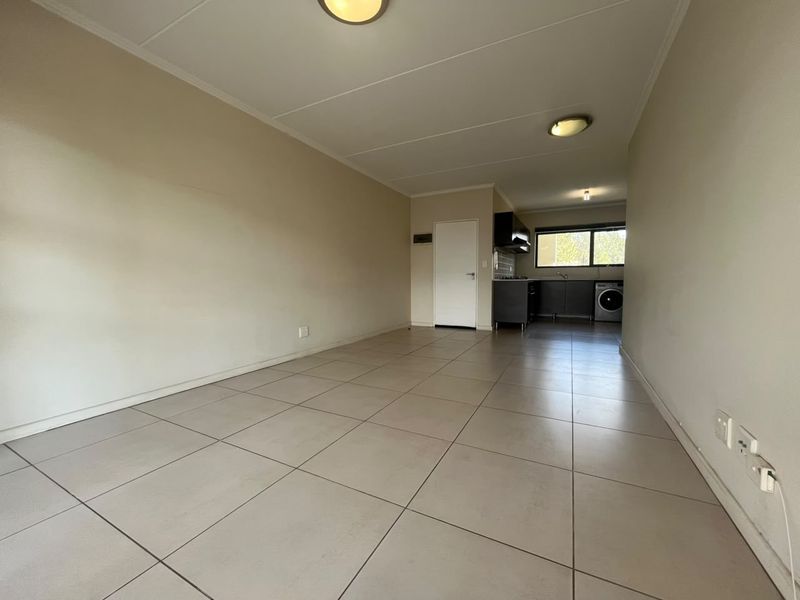 An open-plan living and kitchen area featuring neutral tiled flooring, built-in cabinetry, and integrated kitchen appliances in a clean, vacant space.