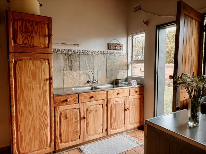 A functional kitchen or scullery area featuring wooden cabinetry, a double stainless steel sink, and natural stone tile backsplash with direct outdoor access.