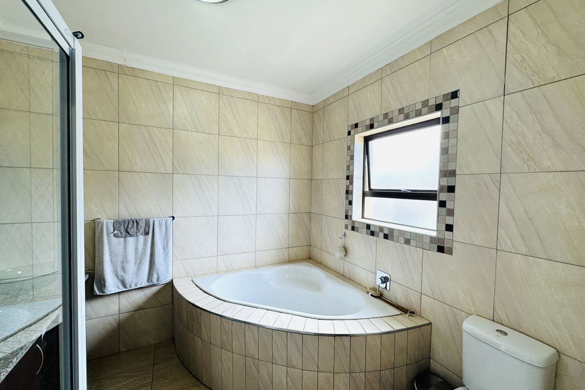 A well-maintained bathroom featuring a corner bathtub, tiled walls and flooring, a shower enclosure, and a top-hung window for natural light.