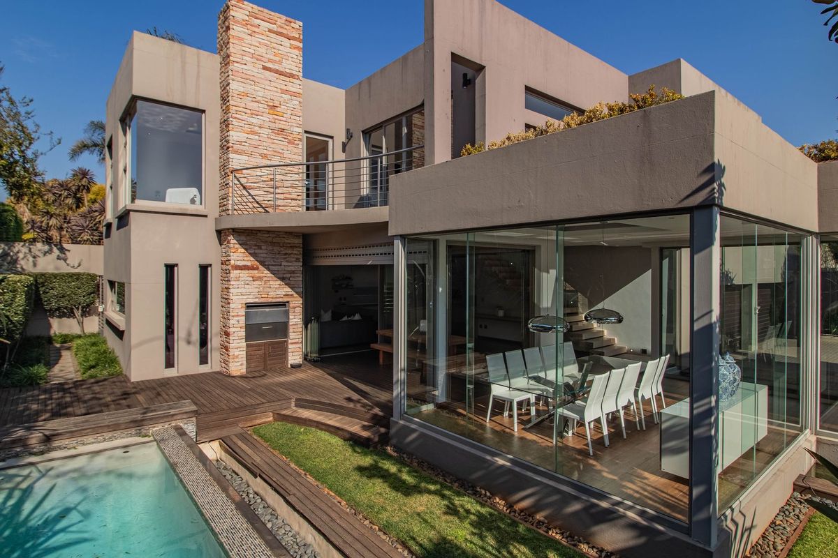 Modern rear elevation featuring a swimming pool, timber decking, stone-clad chimney, and a striking glass-enclosed dining area with an upper-level balcony.