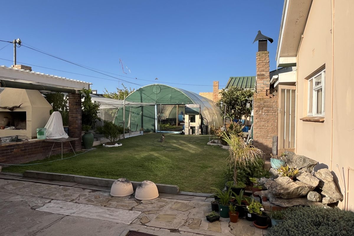 A spacious rear garden featuring a lawn, a large greenhouse structure, a built-in outdoor fireplace, and paved patio area.