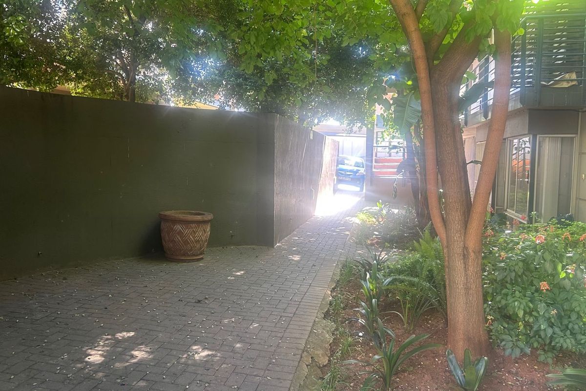 A paved rear walkway featuring a retaining wall and established garden beds, providing a shaded outdoor pathway alongside the townhouse structure.