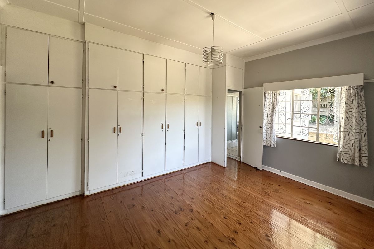 A spacious bedroom featuring extensive built-in cupboards, polished hardwood flooring, and a window with security bars.