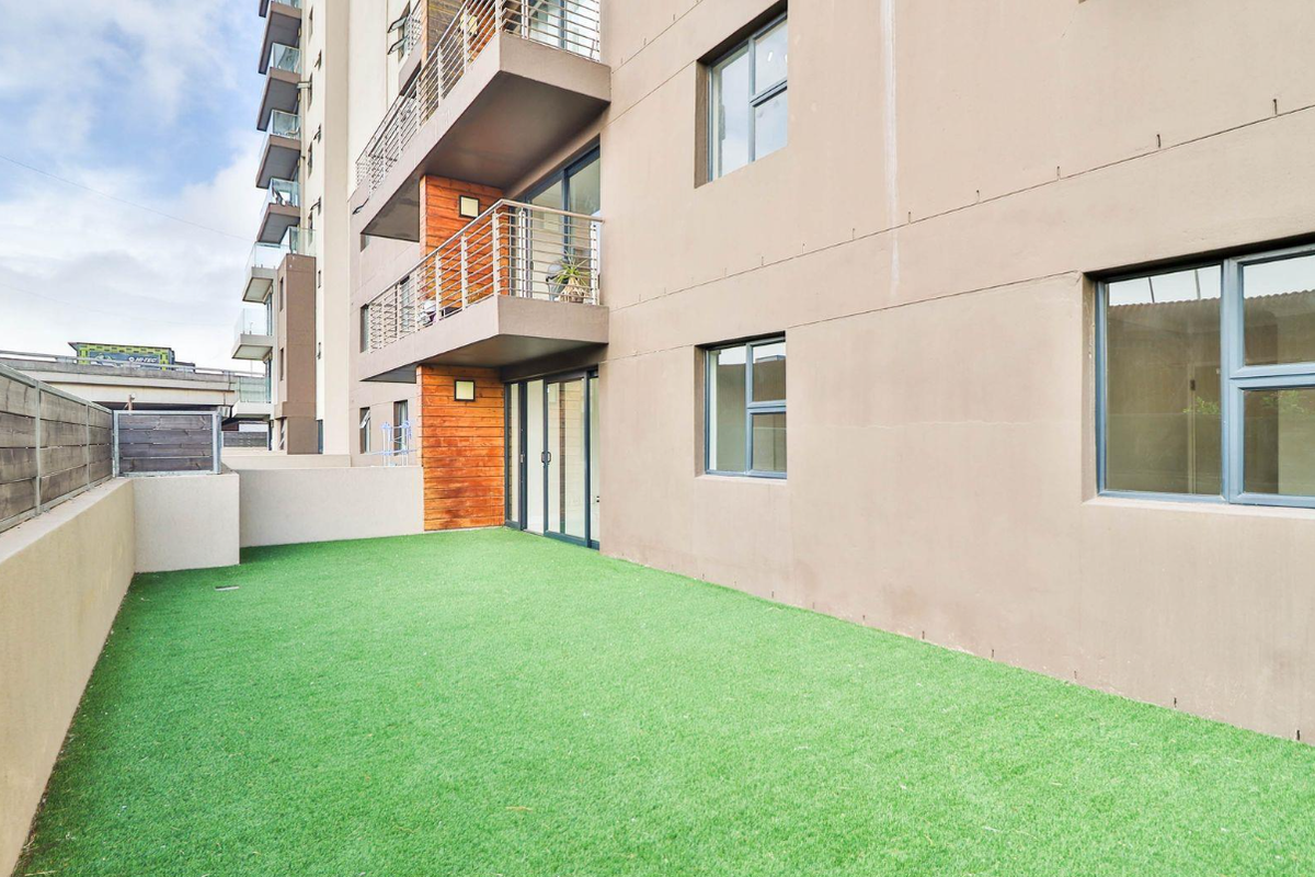 A ground floor apartment exterior featuring a private artificial grass garden area, sliding door access, and modern building facade with balconies.
