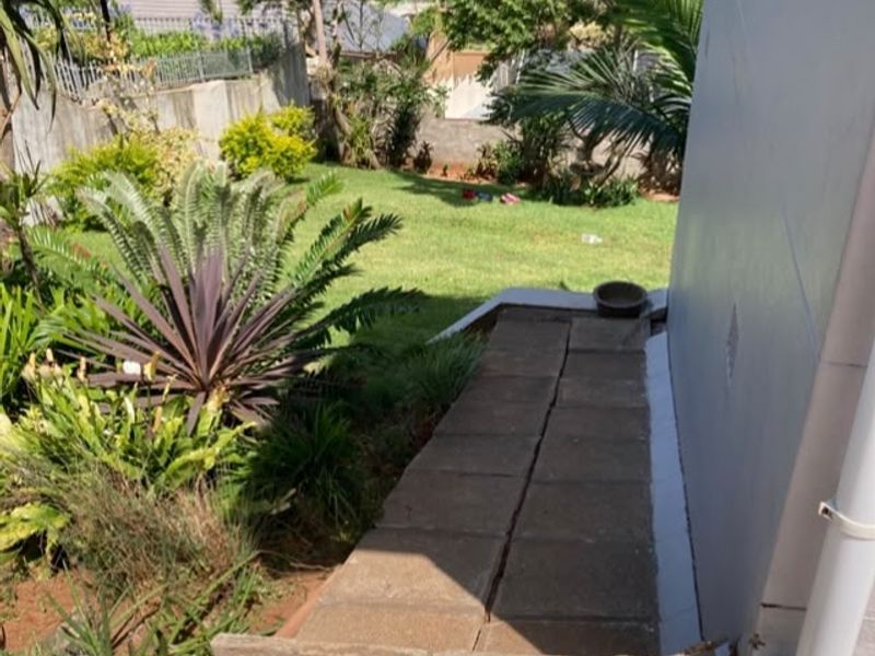 A view of the side garden featuring a paved walkway, landscaped greenery, and a functional downpipe system along the exterior wall.