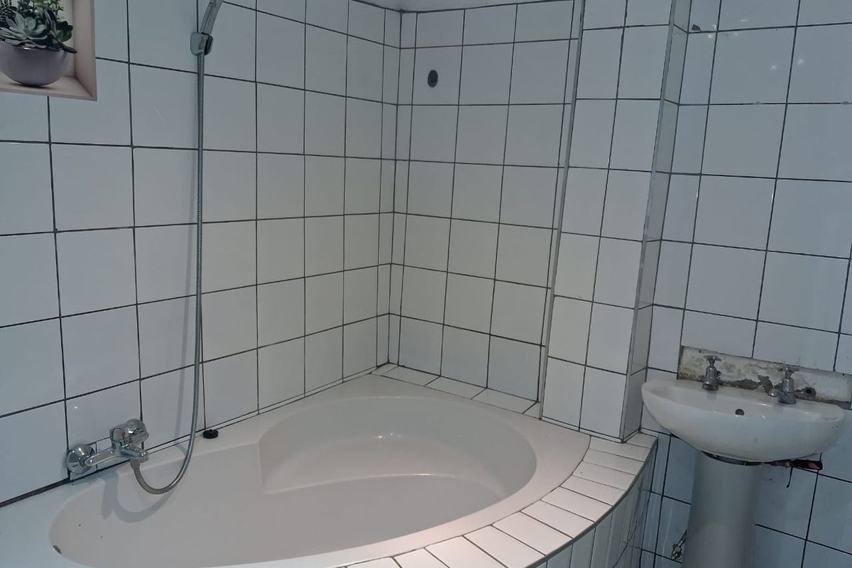 A bathroom featuring a built-in corner bathtub with a shower attachment and a pedestal basin, requiring some cosmetic repairs to the wall tiling.