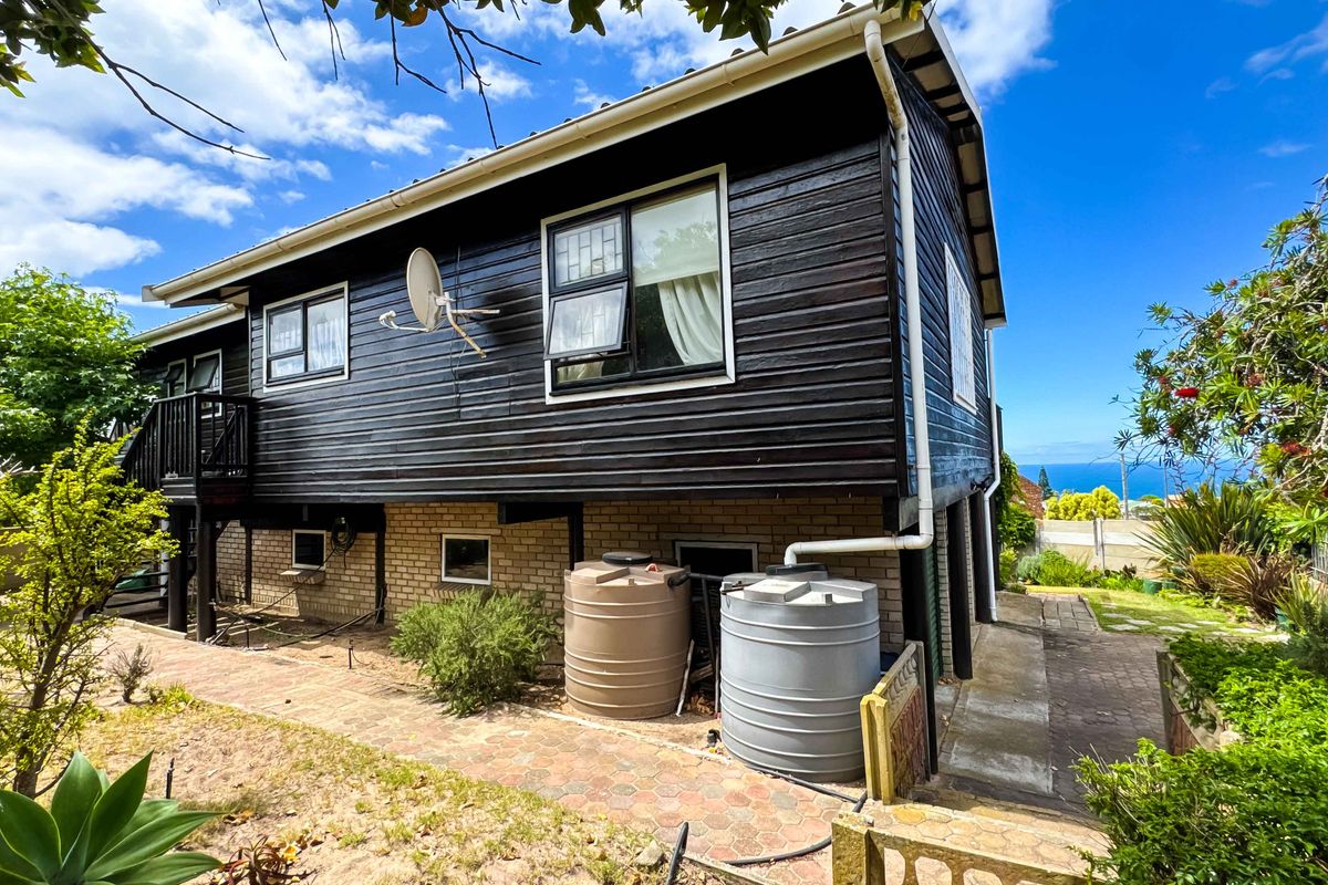 Rear exterior view of a timber-clad house featuring brick foundation, rainwater tanks, and paved patio area with distant ocean views.