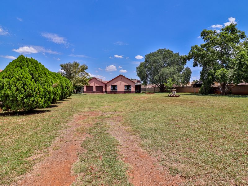 A spacious rear garden featuring a large lawn, mature trees, perimeter walling, and a double garage with a concrete driveway.