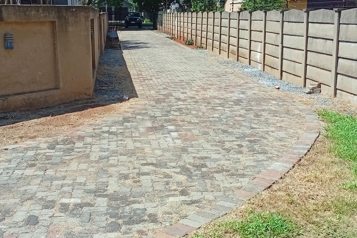 A paved driveway leading towards the rear of the property, enclosed by concrete boundary walls and landscaped garden borders.