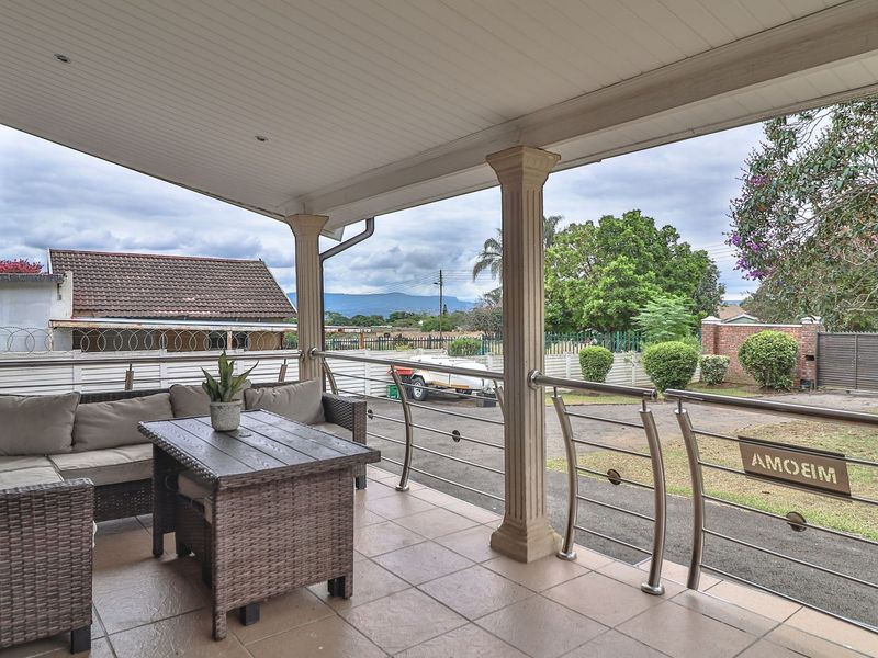 A covered patio featuring tiled flooring, decorative columns, and a stainless steel balustrade overlooking the front driveway and garden area.