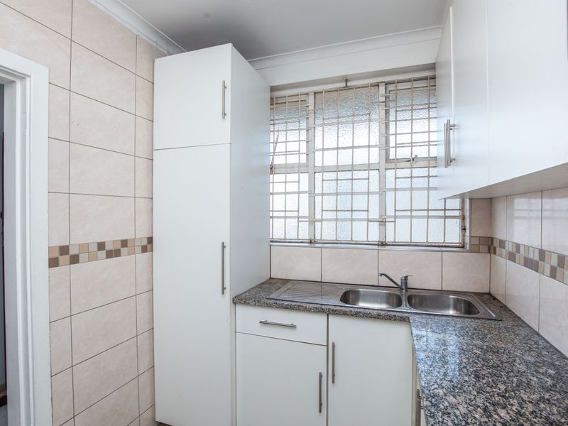 A compact kitchen featuring white built-in cabinetry, granite countertops, a stainless steel double sink, and tiled splashbacks with security bars on the window.