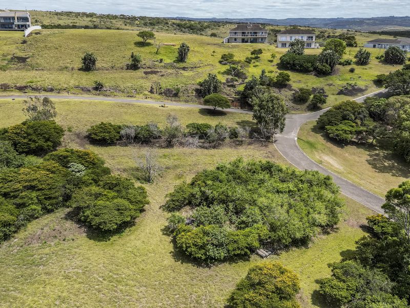 An aerial view of a vacant residential plot featuring sloped terrain, indigenous vegetation, and access to a paved estate road.