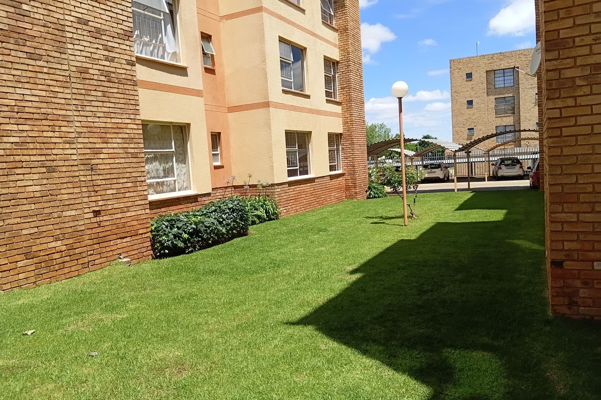 A view of the apartment complex exterior featuring a well-maintained lawn, face brick and plastered facade, and communal parking area with carports.
