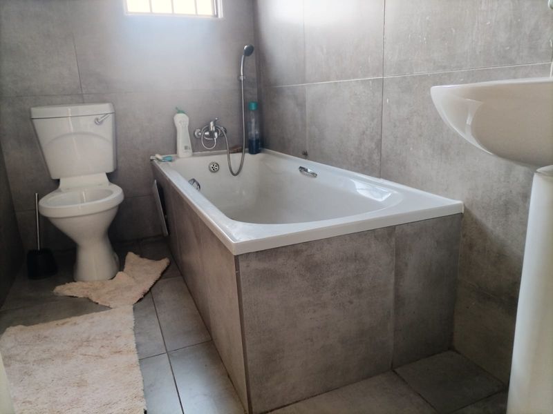A modern bathroom featuring grey tiled walls and flooring, equipped with a standard toilet, a bathtub with a handheld shower mixer, and a pedestal basin.