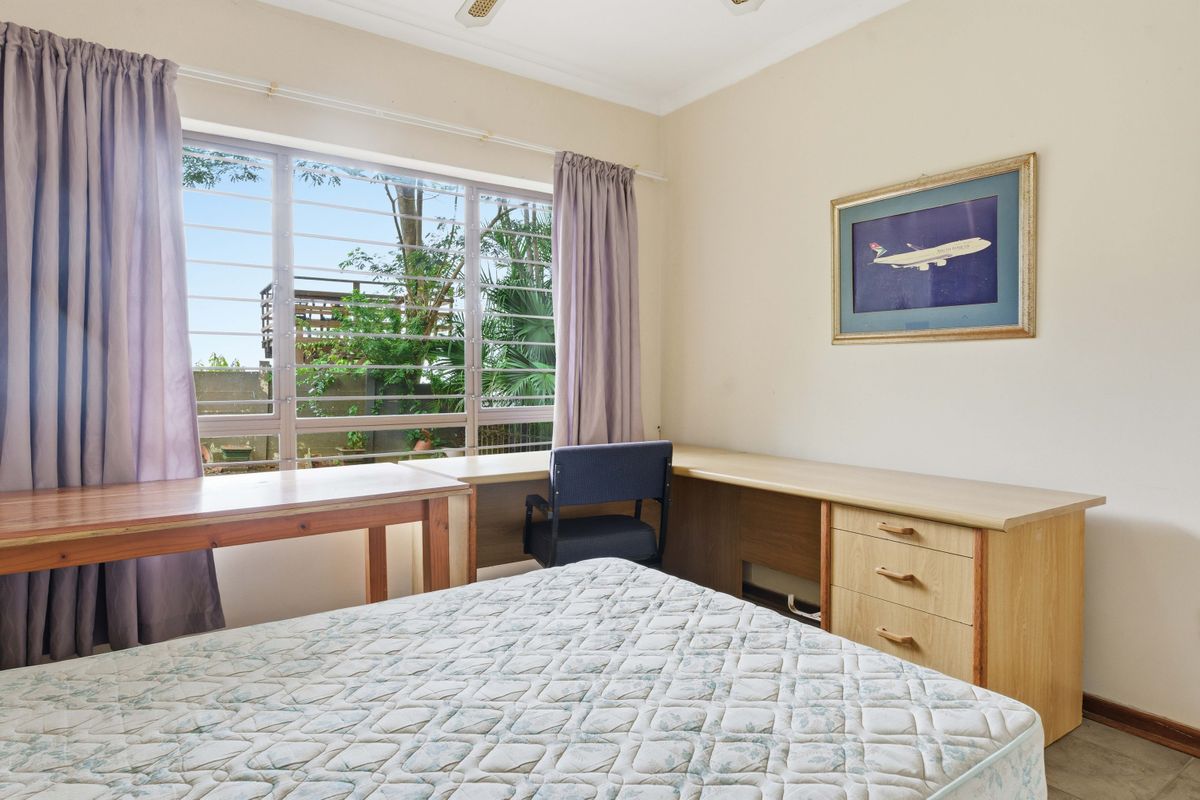A functional bedroom featuring a large window with security bars, built-in wooden desk cabinetry, and neutral wall finishes.
