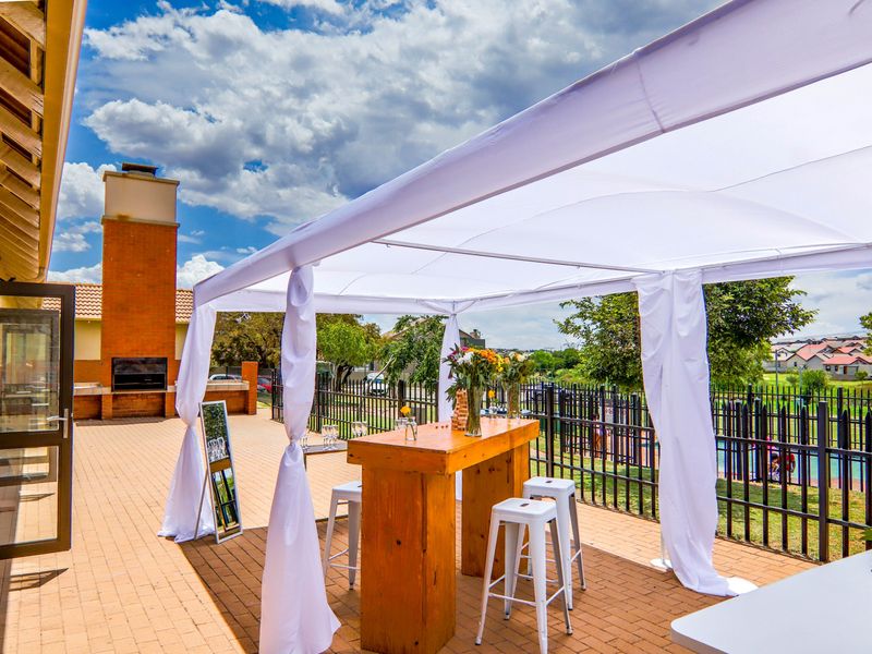 A spacious paved patio area featuring a built-in brick fireplace and covered outdoor space, overlooking a fenced communal area.