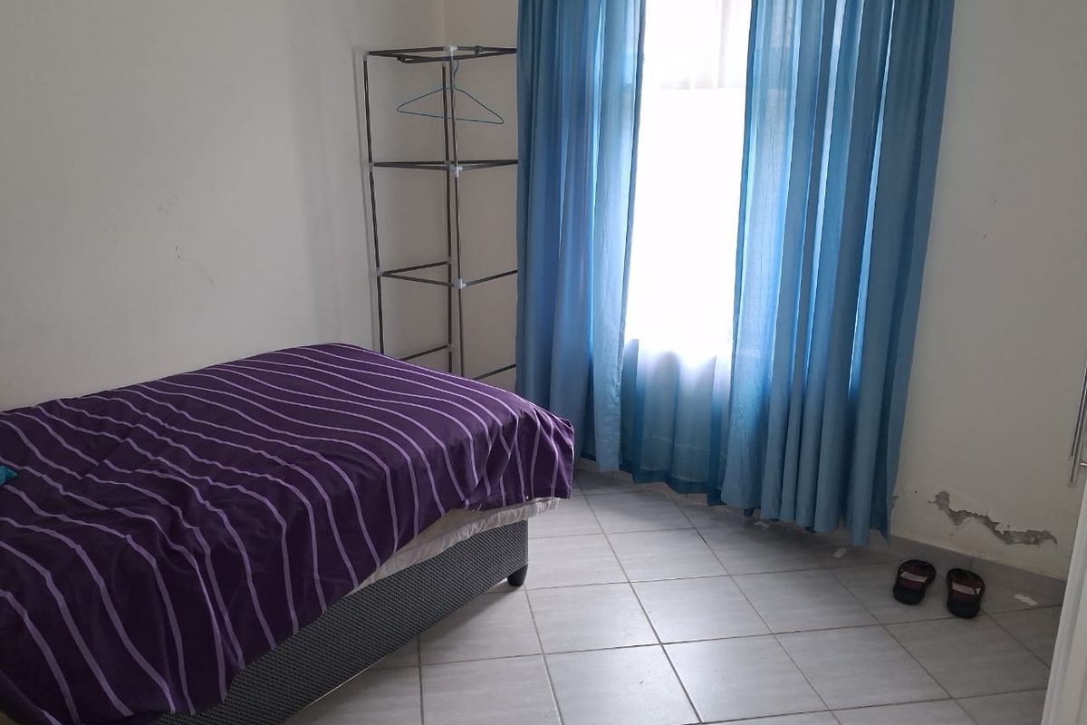 A bedroom featuring tiled flooring and a window with blue curtains. The room shows signs of wall surface deterioration.