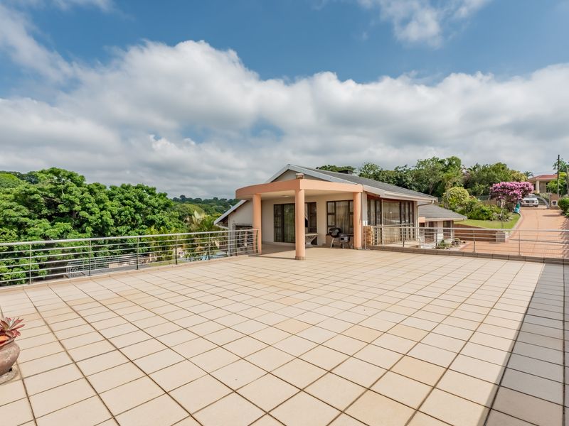 A spacious, tiled outdoor balcony and patio area featuring stainless steel railings, offering an expansive view of the surrounding landscape and property grounds.