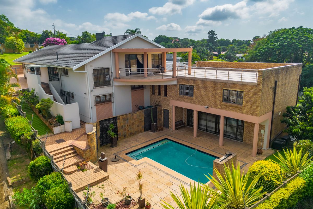 Aerial view of a multi-level residential property featuring a swimming pool, expansive tiled patio area, upper-level balcony, and established garden surroundings.